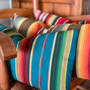 May include: Four colorful striped throw pillows with a southwestern design in shades of orange, red, brown, teal, green, and cream. The pillows are arranged on a wooden chair.