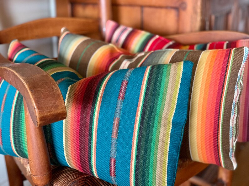 May include: Four colorful striped throw pillows with a southwestern design in shades of orange, red, brown, teal, green, and cream. The pillows are arranged on a wooden chair.