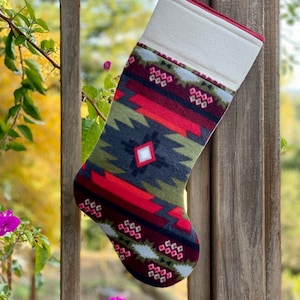 Puede incluir: Una media de Navidad con estampado rojo, verde y negro con un puño blanco. La media está colgada en una barandilla de madera.