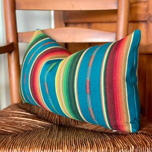 May include: A teal blue and multi-colored striped throw pillow with a woven texture. The pillow is sitting on a woven chair.