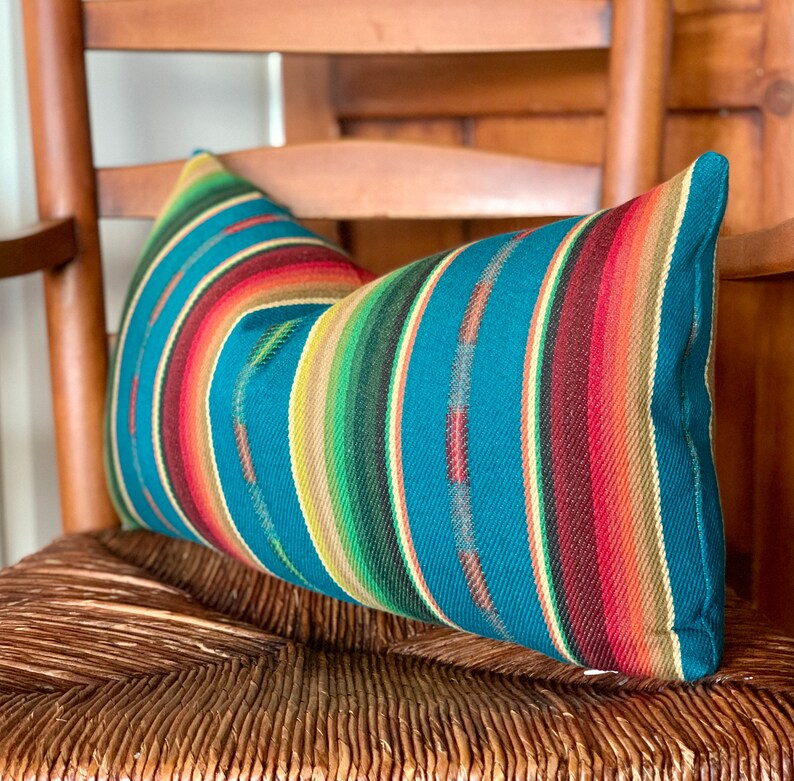 May include: A teal blue and multi-colored striped throw pillow with a woven texture. The pillow is sitting on a woven chair.