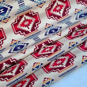 May include: Cream-colored fabric with a repeating geometric pattern in red, blue, and yellow. The design features diamond and triangular shapes, creating a Southwestern or Native American aesthetic. The fabric is folded, showing its texture and drape.