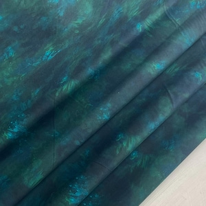 May include: Folded fabric featuring a watercolour design in teal, turquoise, and navy blue. The fabric has a flowing, abstract pattern with lighter and darker tones, creating a textured appearance.