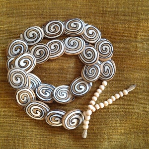 May include: A necklace made of white and brown oval beads with a spiral pattern. The beads are strung on a cord and the necklace is finished with a tassel.