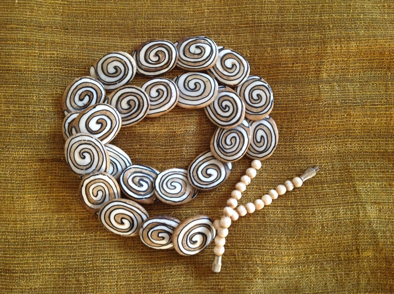 May include: A necklace made of white and brown oval beads with a spiral pattern. The beads are strung on a cord and the necklace is finished with a tassel.