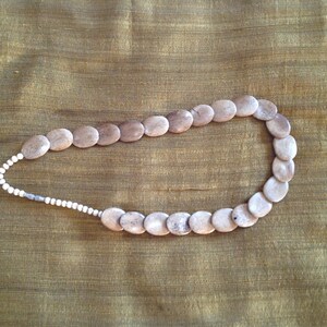 May include: A necklace made of light brown oval-shaped beads with a white bead accent. The necklace is on a brown fabric background.