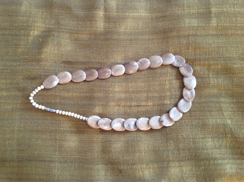 May include: A necklace made of light brown oval-shaped beads with a white bead accent. The necklace is on a brown fabric background.