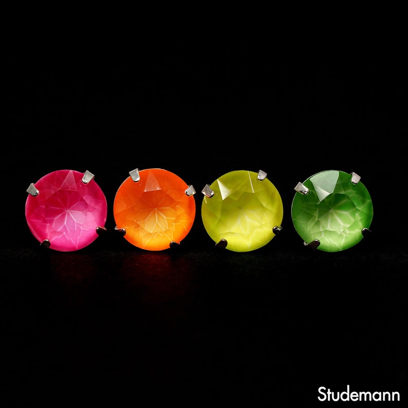 May include: Four colorful gemstone earrings in pink, orange, yellow-green, and green, each with a silver setting. The gems are round and faceted, with a bright, luminous appearance against a black background. The Studemann logo is in the bottom right corner.