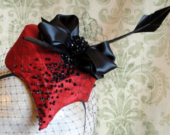 May include: A red fascinator hat featuring a black satin bow, black beaded embellishments, and a black netting veil. A long, thin black arrow-shaped ornament extends from the side of the hat, which is mounted on a thin black headband.