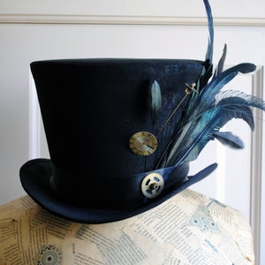 May include: A black top hat with a steampunk aesthetic. The hat features a black ribbon, a small clock-face pin, and a gear-shaped embellishment. Dark blue feathers and a gold pin add further detail to the hat's design.