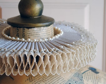 May include: A pleated ruff collar made of beige paper with printed Greek text, featuring a circular row of small white pearls along the inner edge. The collar is topped with a dark metallic sphere and rests on a base covered in newspaper print.
