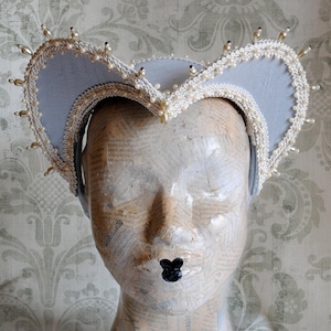 May include: A light blue French hood headpiece featuring a curved, heart-shaped frame edged with intricate white braided trim and small, cream-colored pearl beads spaced along the entire outer perimeter of the structured fabric head covering.