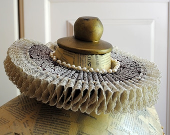 May include: A ruff collar featuring pleated beige lace and dark brown fabric with a printed pattern. The collar is adorned with a string of small white pearls and sits atop a mannequin torso covered in newspaper print with a gold-painted wooden knob.