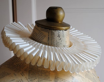 May include: A white pleated ruff collar made of stiffened paper or fabric, featuring a circular design with uniform folds. The collar is displayed around the neck of a mannequin covered in decoupage newspaper print with a gold-colored wooden finial.