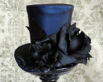 Puede incluir: Un sombrero de copa azul oscuro con una banda de cinta de satén negra, una gran rosa de tela negra y pequeños detalles florales negros. Un fino velo de red negro se extiende desde el ala, colocado sobre un soporte de pedestal negro.