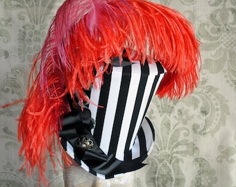 May include: A top hat featuring vertical black and white stripes, adorned with a large cluster of vibrant red and pink ostrich feathers. The base of the crown is decorated with a black ribbon, a dark metallic filigree ornament, and clear gems.