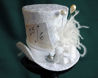 May include: A white top hat featuring a lace overlay, a white satin ribbon band, and a small silver-toned teapot charm. A paper card with the text 10/6 is tucked into the band, accompanied by white fabric flowers, feathers, and two pearl pins.