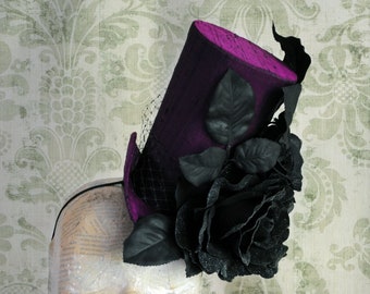 Puede incluir: Un mini sombrero de copa en color morado oscuro con acabado texturizado, adornado con grandes rosas de tela negra, hojas de color verde oscuro y un fino velo de red negra. El sombrero está montado sobre una diadema negra fina y estructurada.
