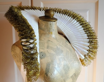 May include: A large, circular, pleated white ruff collar featuring a decorative gold metallic mesh trim along the outer and inner edges. The collar is displayed on a mannequin torso covered in printed paper with various text and graphic elements.