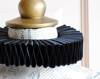 May include: A black pleated ruff collar sits atop a mannequin torso covered in newspaper print. The collar features a circular gold-colored wooden finial at the center. The mannequin is constructed from paper with visible text in various fonts.