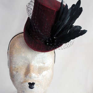 May include: A deep red top hat fascinator featuring a textured fabric finish, adorned with a black netting veil and a cluster of black feathers. The hat is secured with a thin black headband and includes small black decorative beads at the base.