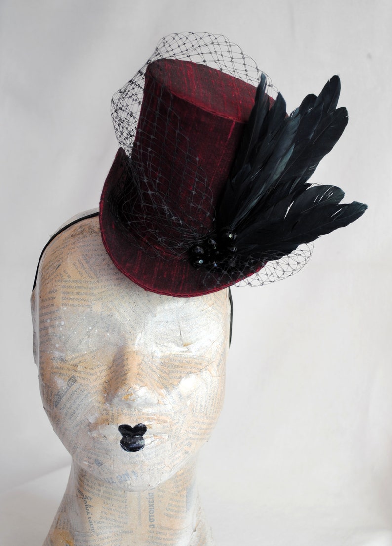 May include: A deep red top hat fascinator featuring a textured fabric finish, adorned with a black netting veil and a cluster of black feathers. The hat is secured with a thin black headband and includes small black decorative beads at the base.