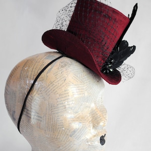 May include: A deep burgundy top hat fascinator attached to a thin black headband. The hat features a black netting veil draped over the crown and brim, accented with black feathers and dark decorative beads on the side of the brim.
