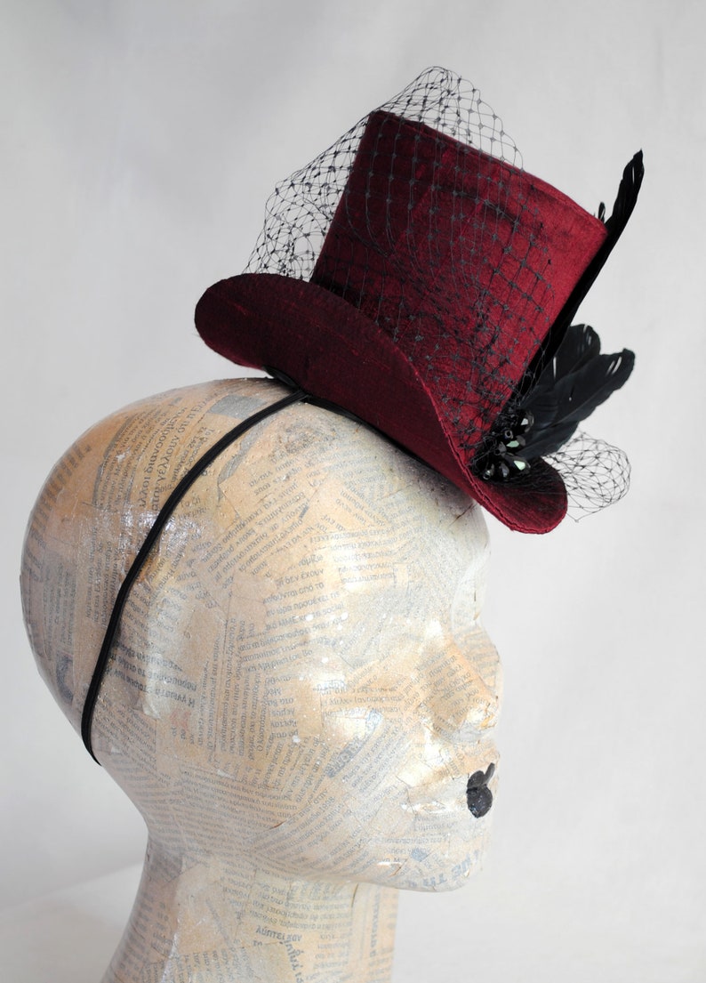 May include: A deep burgundy top hat fascinator attached to a thin black headband. The hat features a black netting veil draped over the crown and brim, accented with black feathers and dark decorative beads on the side of the brim.