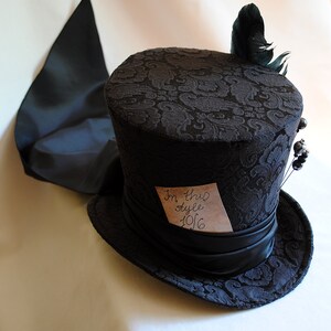 May include: A black top hat with a patterned fabric design and a black satin ribbon. A small piece of paper with the text "In this style 1916" is attached to the hat. A black feather is attached to the top of the hat.