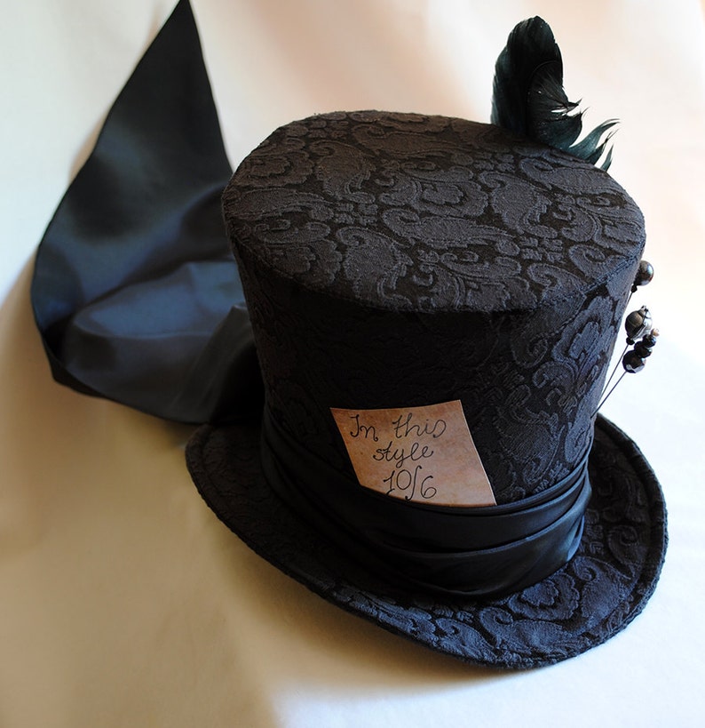 May include: A black top hat with a patterned fabric design and a black satin ribbon. A small piece of paper with the text "In this style 1916" is attached to the hat. A black feather is attached to the top of the hat.