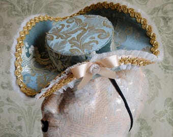 Puede incluir: Un sombrero de ala ancha con un patrón de damasco en azul claro y dorado, adornado con un borde trenzado de metal dorado y encaje blanco con volantes. Un lazo de cinta beige con una perla central decora la parte frontal, unido a una diadema negra.