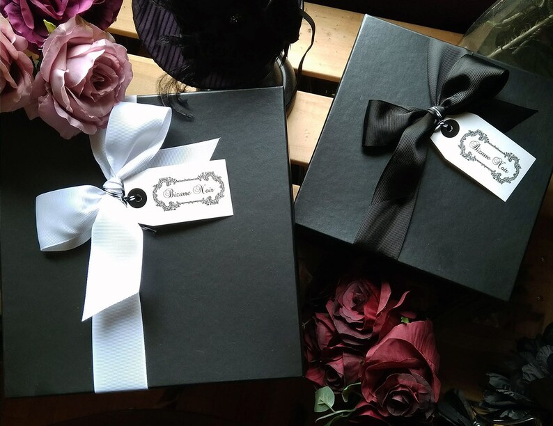 May include: Two black gift boxes with white and black ribbon bows. The boxes are tied with a white ribbon and a black ribbon. Each box has a white tag with the text "Bizane Noir" on it.