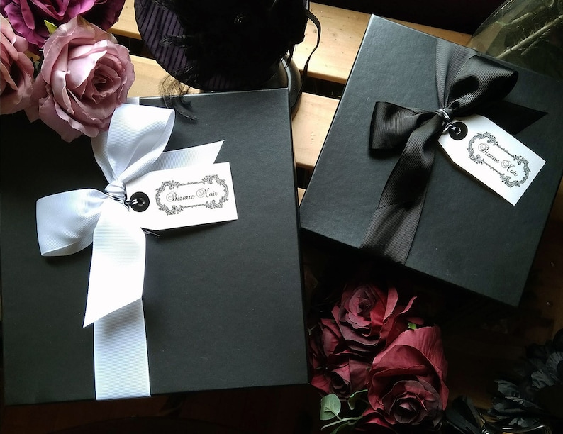 May include: Two square black gift boxes, one tied with a white ribbon and the other with a black ribbon. Each box features a white rectangular tag with the text Bizane Noir in a decorative frame, accented by dark red and purple artificial roses.