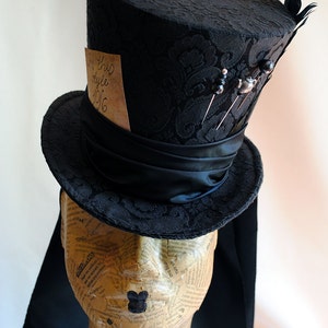 May include: A black top hat with a black satin band and a black feather. The hat is decorated with pins and a small piece of paper with the text "In this style 1916".
