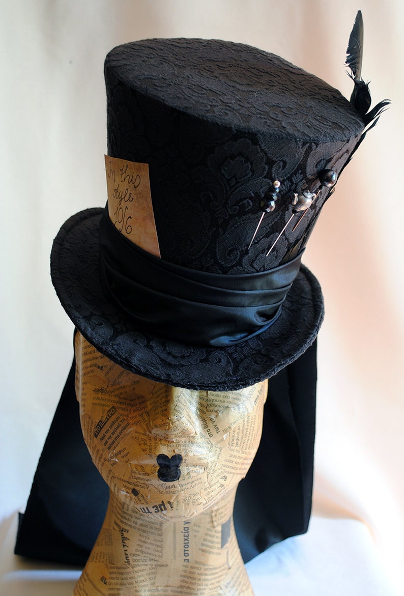 May include: A black top hat with a black satin band and a black feather. The hat is decorated with pins and a small piece of paper with the text "In this style 1916".