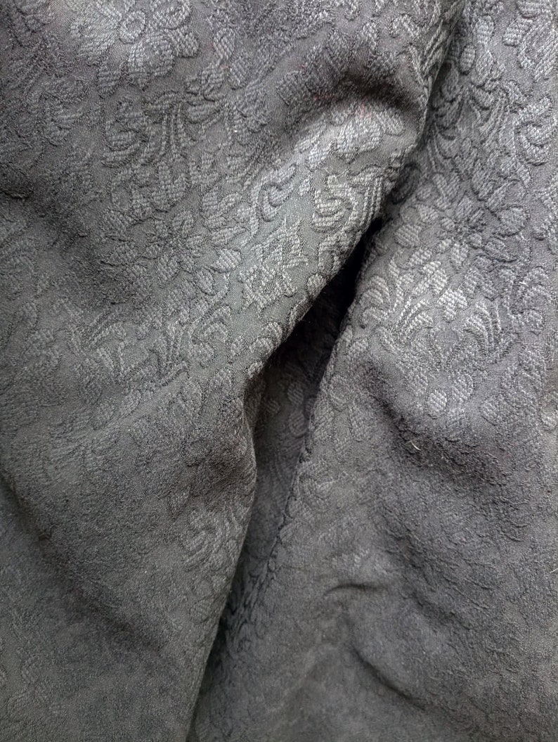 May include: Close-up of a black fabric with a subtle embossed floral pattern. The fabric is wrinkled and folded, showing the texture of the material.