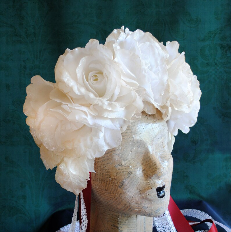 May include: A headpiece featuring a cluster of large, white artificial roses with subtle glitter accents. The floral arrangement is mounted on a mannequin head covered in vintage-style printed paper, with red ribbons and white lace trim hanging down.