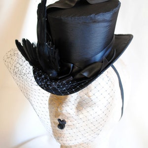 May include: A black top hat with a black feather and a black veil. The hat is sitting on a mannequin head.
