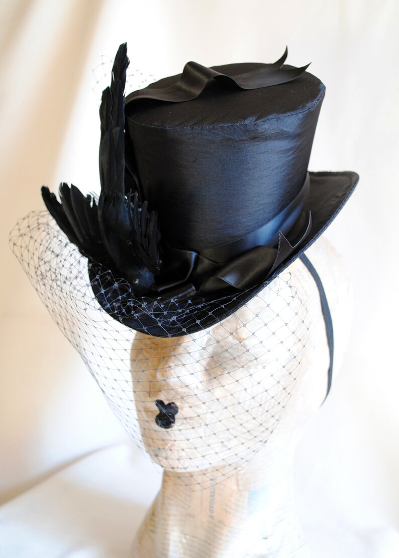 May include: A black top hat with a black feather and a black veil. The hat is sitting on a mannequin head.