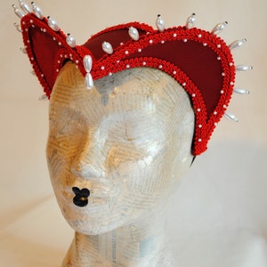 May include: A deep red fabric headpiece featuring a structured, curved silhouette adorned with intricate red braided trim along the edges. The headpiece is embellished with a series of white, teardrop-shaped faux pearls spaced evenly along the outer rim.