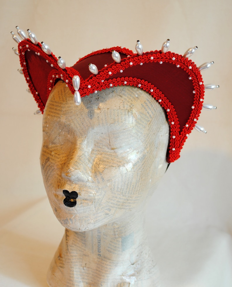 May include: A deep red fabric headpiece featuring a structured, curved silhouette adorned with intricate red braided trim along the edges. The headpiece is embellished with a series of white, teardrop-shaped faux pearls spaced evenly along the outer rim.