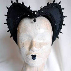 May include: A black fabric headpiece featuring a heart-shaped silhouette with intricate braided trim along the edges. The headpiece is adorned with small black beads along the perimeter and a central faceted black gemstone with a dangling teardrop bead.