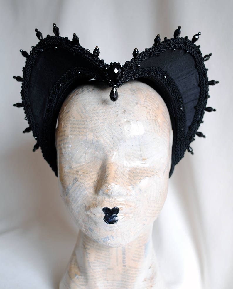 May include: A black fabric headpiece featuring a heart-shaped silhouette with intricate braided trim along the edges. The headpiece is adorned with small black beads along the perimeter and a central faceted black gemstone with a dangling teardrop bead.