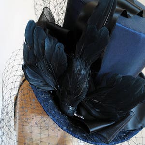May include: A blue top hat with a black velvet band and a black feather embellishment. The hat is adorned with a black bird feather and a black veil.
