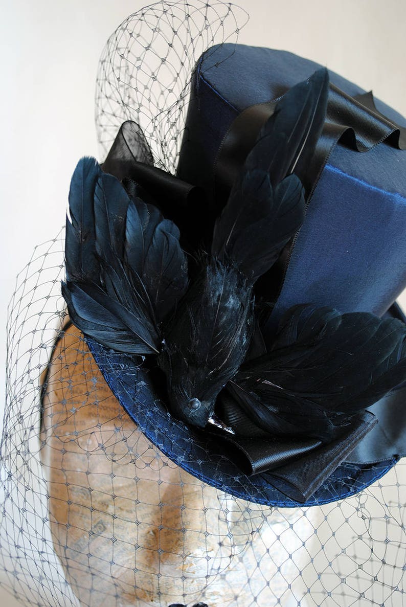 May include: A blue top hat with a black velvet band and a black feather embellishment. The hat is adorned with a black bird feather and a black veil.