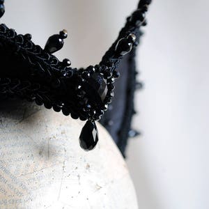May include: A black crown featuring intricate braided trim, faceted black beads, and dangling teardrop-shaped crystals. The headpiece is constructed with a structured base and adorned with multiple small, dark, reflective bead accents throughout.