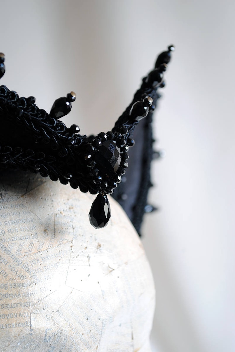 May include: A black crown featuring intricate braided trim, faceted black beads, and dangling teardrop-shaped crystals. The headpiece is constructed with a structured base and adorned with multiple small, dark, reflective bead accents throughout.