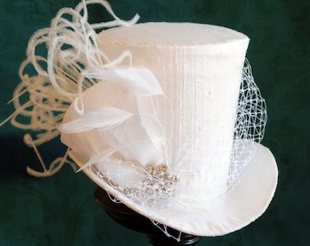 May include: A white top hat featuring a textured fabric finish, adorned with white feathers, a delicate white netting veil, and a central silver-toned crystal embellishment positioned on the side of the brim for a detailed decorative appearance.