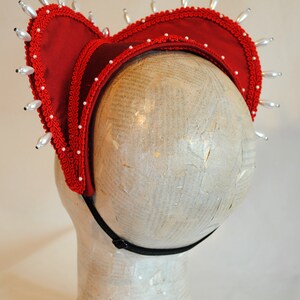 May include: A red fabric headpiece featuring a structured heart-shaped frame, adorned with white teardrop-shaped beads along the outer edge and smaller white beads along the inner trim. The piece includes a black elastic strap for secure placement.