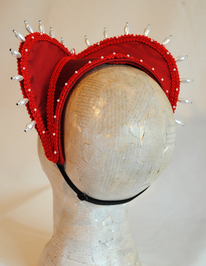 May include: A red fabric headpiece featuring a structured heart-shaped frame, adorned with white teardrop-shaped beads along the outer edge and smaller white beads along the inner trim. The piece includes a black elastic strap for secure placement.